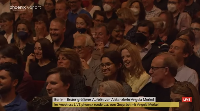 Зрители, пришедшие послушать бывшего канцлера, смеются над рассказами Меркель про Владимира Путина. Фото: скрин видео You Tube